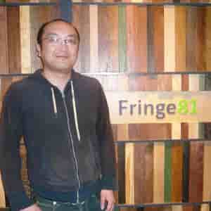 Fringe81株式会社のロゴを背景に立つ田中弦氏の写真